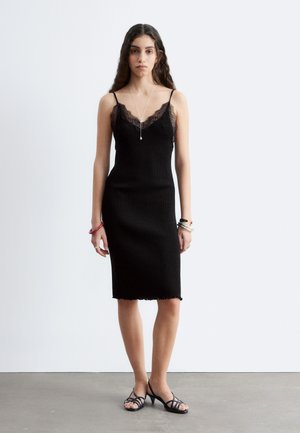 Mujer de pie con vestido negro acanalado con encaje, sandalias de tacón alto, collar largo y varias pulseras contra una pared blanca lisa.