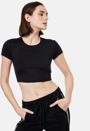 Ung kvinde iført en sort cropped T-shirt og sorte træningsbukser med hvide striber, poserende med hænderne i lommerne mod en ensfarvet baggrund.