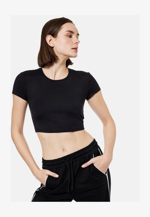 Ung kvinde iført en sort cropped T-shirt og sorte træningsbukser med hvide striber, poserende med hænderne i lommerne mod en ensfarvet baggrund.