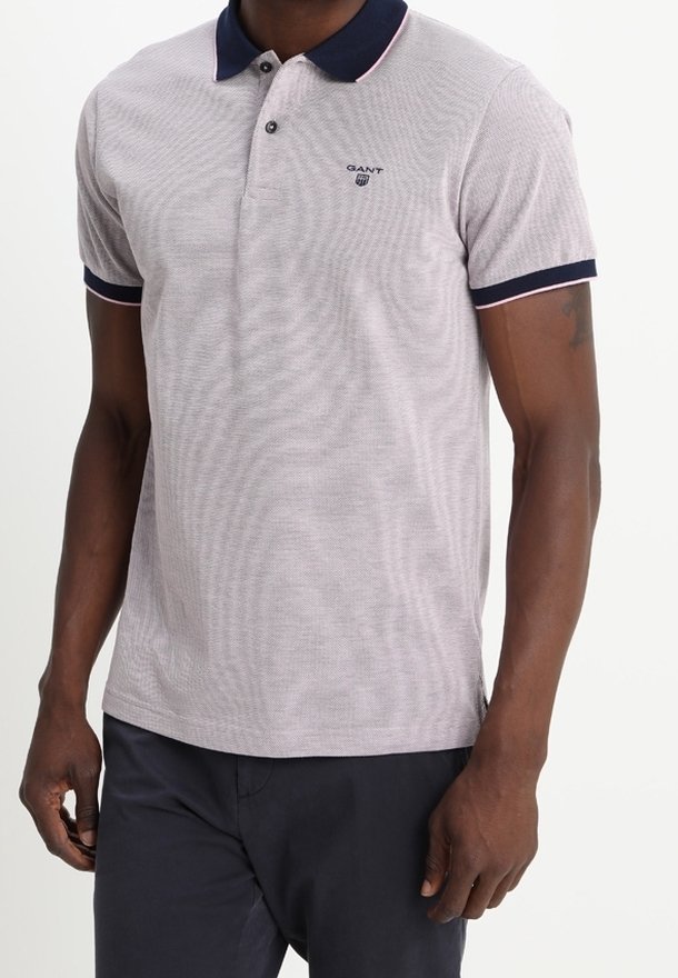 Homme portant un polo gris clair avec col et poignets bleu marine, associé à un pantalon foncé, debout devant un fond uni.