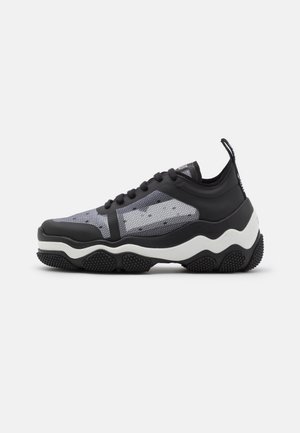 Zapatilla negra y gris con parte superior de malla, cordones negros, suela gruesa blanca y negra con ondas y cinta para tirar en el talón, mostrada en vista lateral.