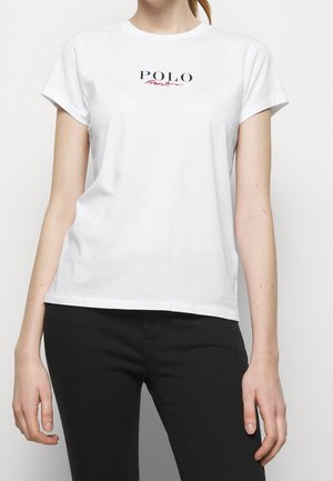 Kvinde iført en hvid kortærmet t-shirt med teksten "POLO" og rød kursiv skrift på brystet, kombineret med sorte bukser.