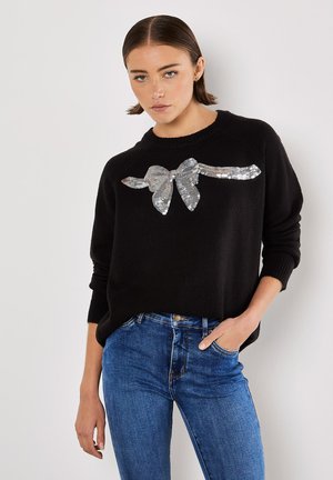 Pull en maille noire avec un col rond et un motif de nœud en sequins argentés sur le devant, associé à un jean en denim bleu.