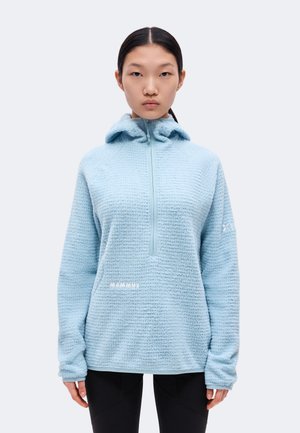 Un mannequin portant un pull polaire bleu clair à capuche et un legging noir, debout devant un fond uni et clair.