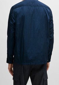 Mörkblå lättviktsjacka med en slät yta, något skrynklig finish och en knappad krage. Har långa ärmar och en avslappnad passform.