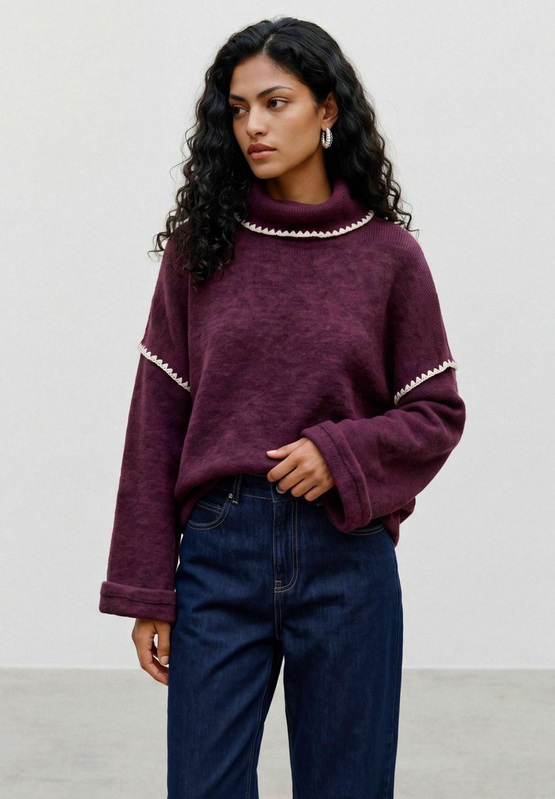 Femme aux cheveux bouclés portant un pull à col roulé violet avec un bord blanc et un jean bleu foncé, debout devant un fond uni.