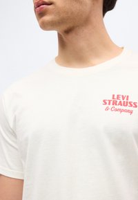 Νέος άνδρας που φοράει λευκό μπλουζάκι με στρογγυλή λαιμόκοψη και κόκκινο κείμενο «Levi Strauss & Company» στο αριστερό στήθος.