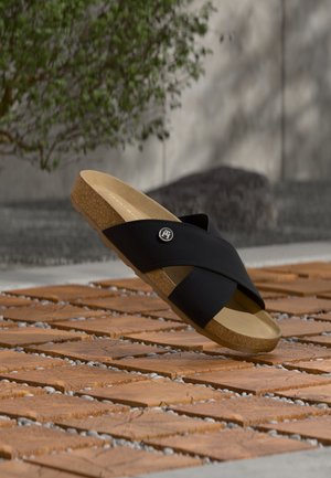 Schwarze Leder-Slidesandale mit überkreuzten Riemen und Korksohle, die tagsüber über Terrakottaplättchen im Freien schwebt.