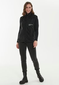 Musta fleece-liivi vetoketjulla, jossa on korkea kaulus ja logo vasemmassa rinnassa, yhdistettynä tummiin farkkuihin ja nilkkabootseihin.