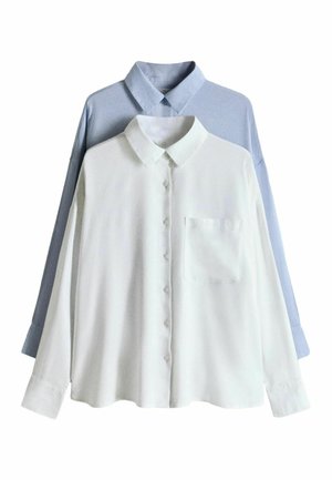 Kobieta w luźnej, jasnoniebieskiej koszuli na guziki z podwiniętymi rękawami, dopasowanych spodniach oraz długim naszyjniku z okrągłym wisiorkiem, patrząca w dół.