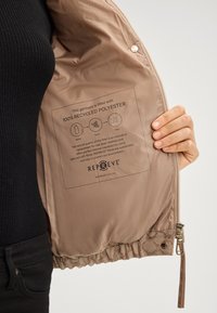 Brązowa kurtka z gładką, błyszczącą podszewką, z metką wskazującą, że jest wypełniona w 100% z recyklingowanego poliestru. Zamek błyskawiczny i elastyczny dół w zestawie.