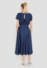 Laivansinis midi-mekko lyhyillä hihoilla, avaimenreikäyksityiskohdalla takana, vyötäisiltä kiristetty ja laskeutuva hame. Yhdistetty hopeisiin korkokenkiin.