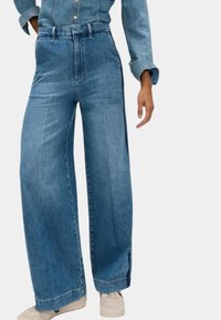 Jeans in denim blu a gamba larga con vita alta, dotati di cuciture laterali e una texture leggermente sfilacciata, abbinati a sneakers di colore chiaro.