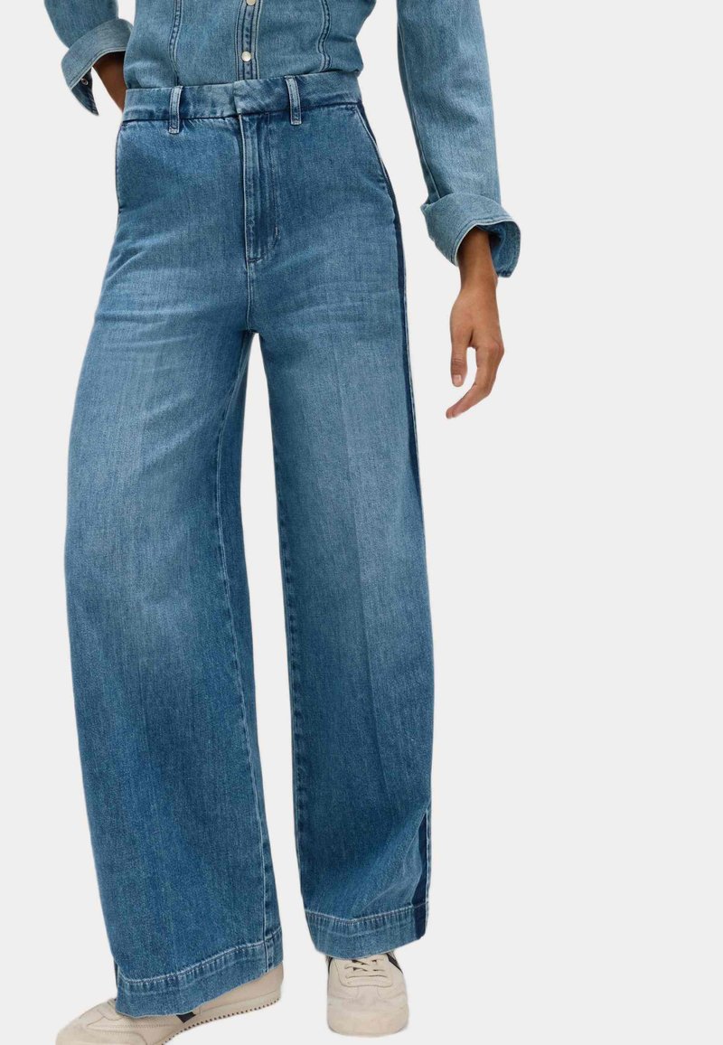 Jeans in denim blu a gamba larga con vita alta, dotati di cuciture laterali e una texture leggermente sfilacciata, abbinati a sneakers di colore chiaro.