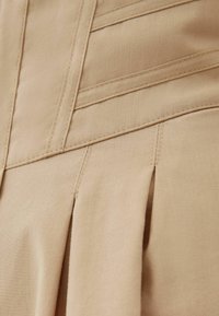 Beige figurnära klänning med texturerad tyg, vertikala sömmar och plisserade detaljer i botten. Slät yta med väldefinierade sömnadsdetaljer.