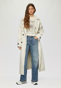 Lichtgrijze lange jas met zwarte knopen, gedragen over een graphic sweatshirt en wijde blauwe jeans, gecombineerd met zwart-witte sneakers.