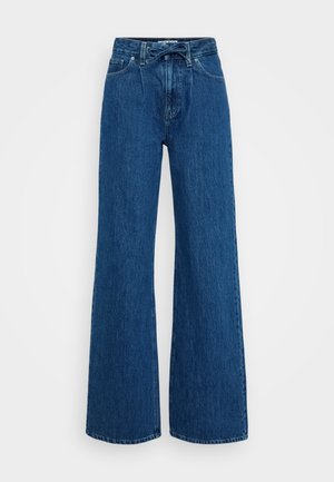 Jeans de mezclilla azul de talle alto con piernas anchas, bolsillos frontales, presillas para cinturón y un cinturón de tela atado a la cintura sobre un fondo simple.