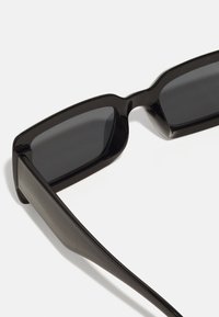 Lunettes de soleil noires avec une épaisse monture rectangulaire, des verres sombres et une finition lisse et brillante sur les branches. Design simple et moderne.