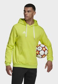 Limettengrüner Hoodie mit Kängurutasche, weißen Kordeln und Adidas-Logo. Das Model hält einen gemusterten Fußball. Schwarze Hose.
