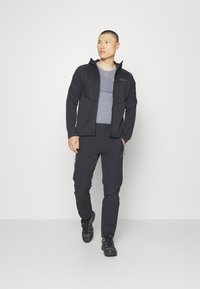 Musta ulkotakki vetoketjulla, säädettävällä hupulla ja harmaalla pitkähihaisella paidalla. Yhdistetty mustiin housuihin ja tukeviin mustiin kenkiin.