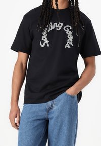 Mustan puuvillan t-paita, jossa on kaarevalla tyylillä "Sporting Goods" -teksti. Lyhyet hihat, rento malli, käytetään vaaleansinisten farkkujen kanssa.