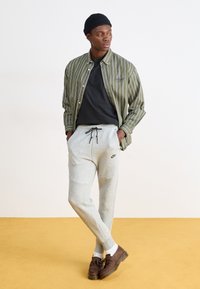 Zeleně pruhovaná košile přes černé tričko, doplněná šedými tepláky s logem Nike a hnědými botami s třásněmi. Žluté pozadí.