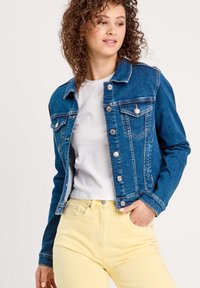 Chaqueta de mezclilla azul con cuello clásico, cierres de botón y dos bolsillos en el pecho, combinada con una camisa blanca y pantalones amarillos de talle alto.