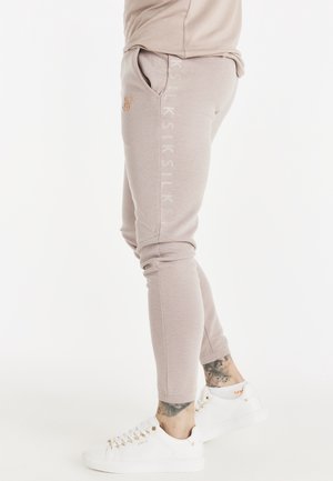 Beige teksturerede joggers med sidelommer, broderet logo nær lommen, brugt med hvide sneakers og synlige ankel-tatoveringer på en lys baggrund.