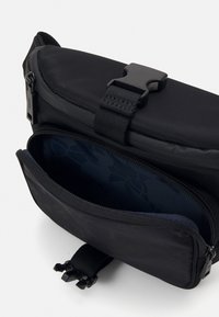 Črn nylon torbica čez telo z odstranljivim sponko. Ima glavni predel in sprednji žep na zadrgo z vzorčasto oblogo.