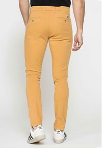 Pantaloni slim-fit giallo senape con due tasche posteriori e una texture liscia, abbinati a scarpe da ginnastica bianche con strisce nere.