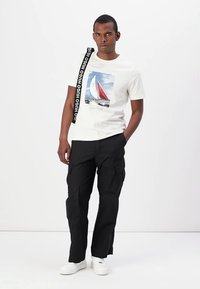 Camiseta de color claro con un gráfico de navegación, combinada con pantalones cargo negros y zapatillas blancas. Presenta una correa negra con acentos de logo.