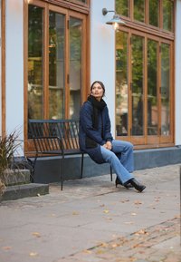Quiltet marineblå frakke med et sort tørklæde, parret med lyseblå flagrende jeans og sorte højhælede støvler, siddende på en sort metalbænk.