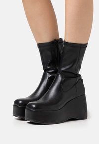 Bottes compensées noires montant jusqu'aux chevilles, en cuir lisse. Dotées d'une fermeture éclair latérale et d'un bout rond, avec une semelle épaisse à plateforme.