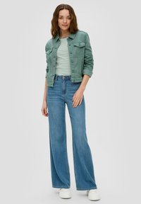 Grüne Jeansjacke über einem gestreiften weißen und grünen Oberteil, kombiniert mit weit geschnittenen blauen Jeans und weißen Sneakern. Glatte Textur, lässiger Stil.