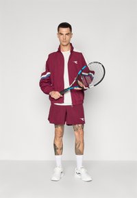 Chaqueta atlética color burdeos con acentos en blanco y azul, combinada con pantalones cortos a juego. Sosteniendo una raqueta de tenis, lleva zapatillas y calcetines blancos.