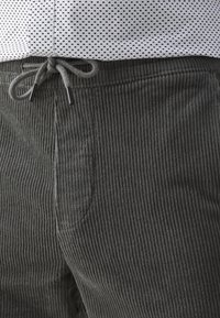 Pantalon en velours côtelé gris avec une taille élastique et un cordon noué en nœud papillon, associé à une chemise blanche ornée d'un petit motif à pois noirs.