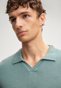 Polo a maniche corte, di un morbido colore teal chiaro, con scollo a V e colletto. Il tessuto ha una texture soffice e presenta lievi dettagli a coste sul collo.