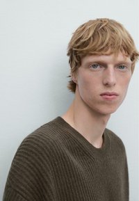Brauner gerippter Pullover mit einer Strickstruktur, der über einen runden Halsausschnitt und einen lässigen Schnitt verfügt. Das Model hat kurzes, hellbraunes Haar und einen neutralen Hintergrund.