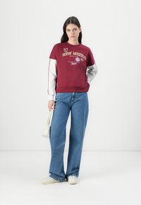 Bordeaux sweatshirt met grijze mouwen en een geribde zoom, met de opdruk "NEW MEXICO SPORTS CLUB". Gecombineerd met een wijdvallende blauwe spijkerbroek en witte schoenen.
