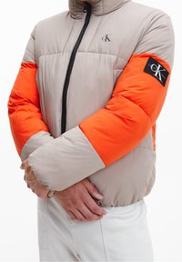 Puhovka v béžové a oranžové barvě, s předním zipem, vysokým límcem a logem na rukávu. Měkká, texturovaná tkanina s návrhem v barevných blocích.