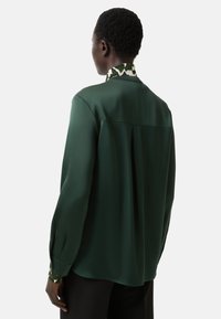 Dunkelgrüne Seidenbluse mit gemustertem Kragen, langen Ärmeln und einer Falte im Rücken für mehr Bewegungsfreiheit. Glatte Textur und taillierte Passform.