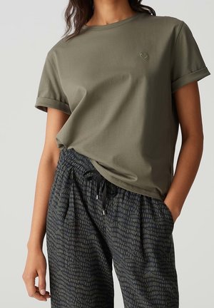 Frau trägt ein olivgrünes kurzärmeliges T-Shirt mit einem kleinen Herz-Emblem und gemusterte Jogginghosen mit Kordelzug und Taschen.