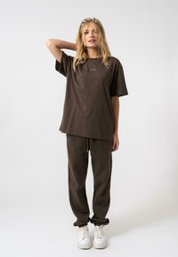 Barna pamut póló rövid ujjakkal és kerek nyakkivágással, hozzá illő sportnadrággal párosítva. Fehér sneakers, texturált talppal. Minimális logó részlet.