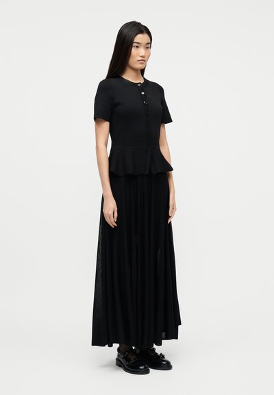 Γυναίκα με μακριά σκούρα μαλλιά, που φοράει μαύρο κοντομάνικο τοπ με κουμπιά και πεπλούμ, καθώς και μακριά μαύρη πλισέ φούστα, στέκεται σε λευκό φόντο.