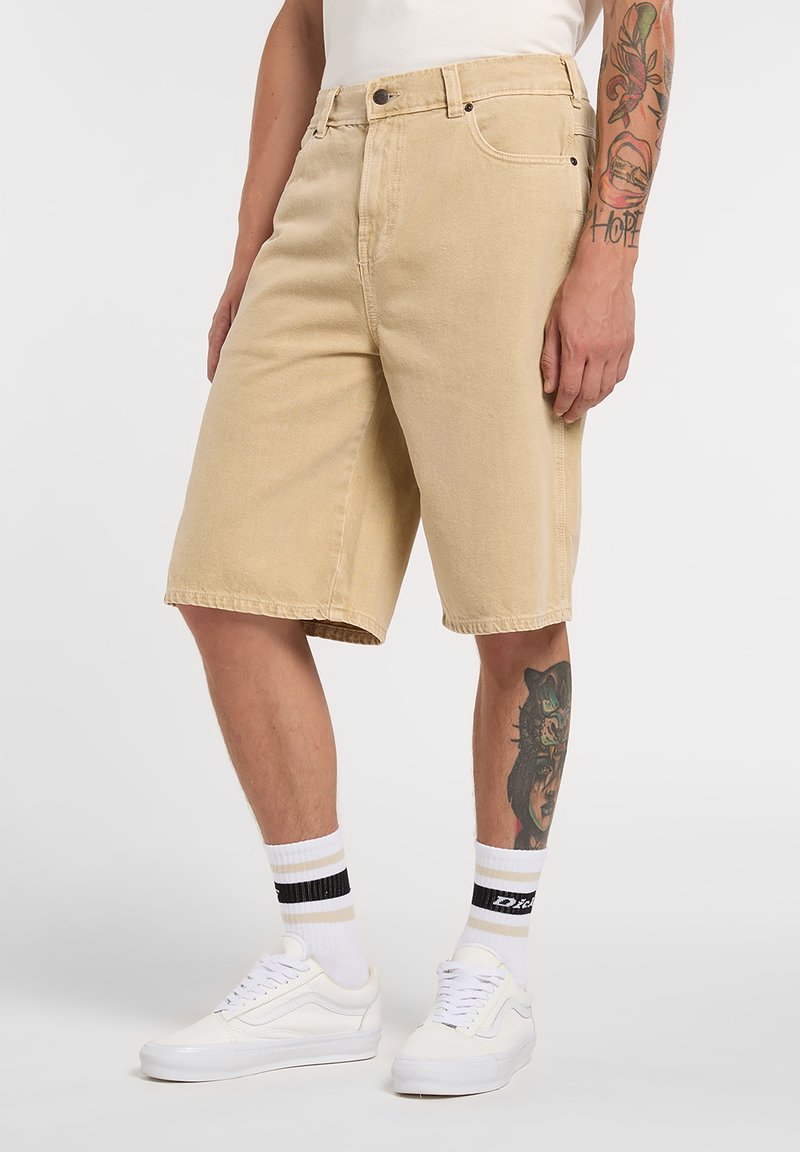 Pantalones cortos de mezclilla beige, por encima de la rodilla, corte clásico, cinco bolsillos, textura sutil, combinados con zapatillas blancas y calcetines a rayas.
