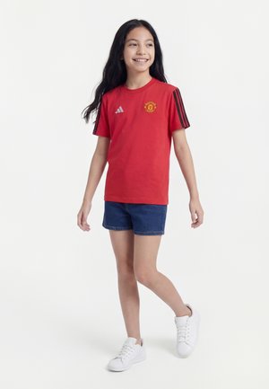 Fille souriante aux longs cheveux noirs portant un t-shirt rouge de Manchester United, un short en jean bleu et des baskets blanches, se tenant sur un fond blanc.