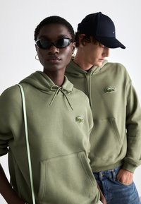 Sudaderas verde olivo con bolsillos tipo canguro y capuchas con cordón, con logotipos de cocodrilo bordados. Una persona lleva gafas de sol negras.