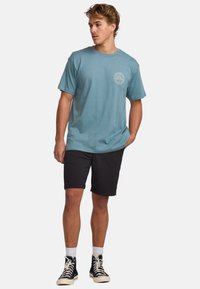 Light blue cotton t-shirt with a round neckline and small logo print on the chest, paired with black shorts and black high-top sneakers.