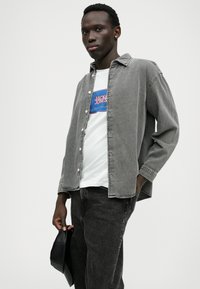 Camisa de denim cinza sobre uma t-shirt branca com um logótipo azul e vermelho, combinada com calças cinza escuras. Segurando uma mala preta. Fundo simples.