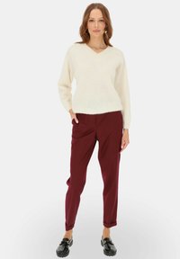 Kremowy, puszysty sweter typu v-neck, luźny krój, w połączeniu z bordowymi spodniami taperowanymi. Model ma na sobie czarne loafersy, co podkreśla elegancki wygląd.